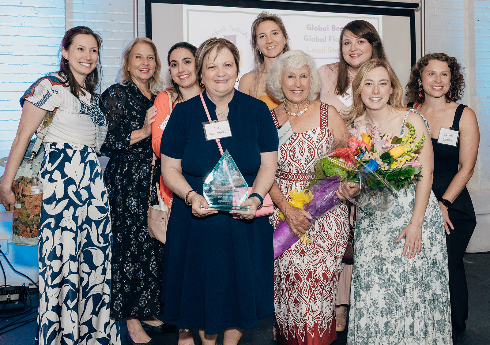 Several alumnae members wearing colorful dresses standing on stage around a member holding an award and bouquets of flowers