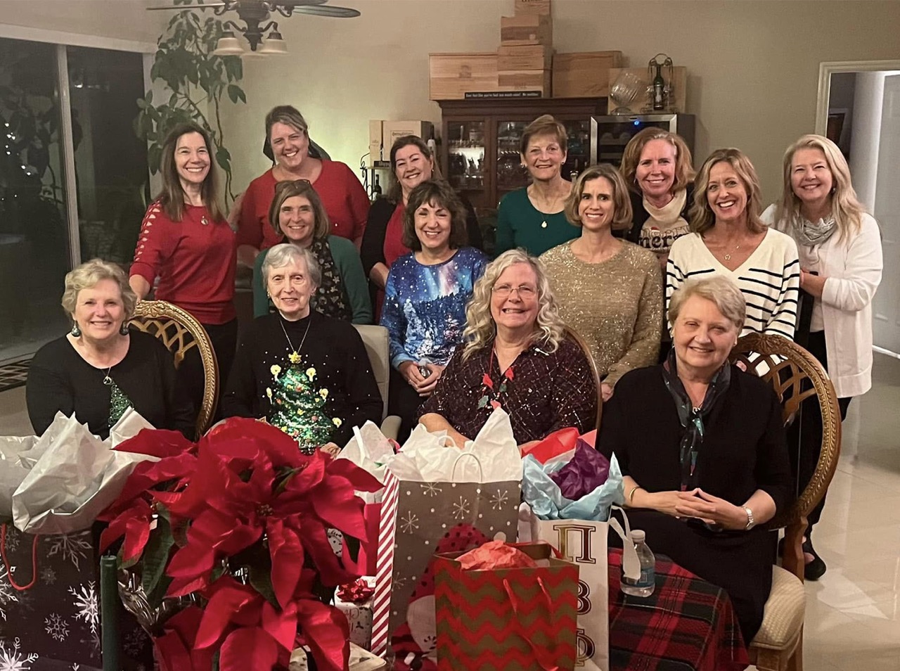 Image of a large group of individuals sitting indoors around a table of festive wrapped gifts.