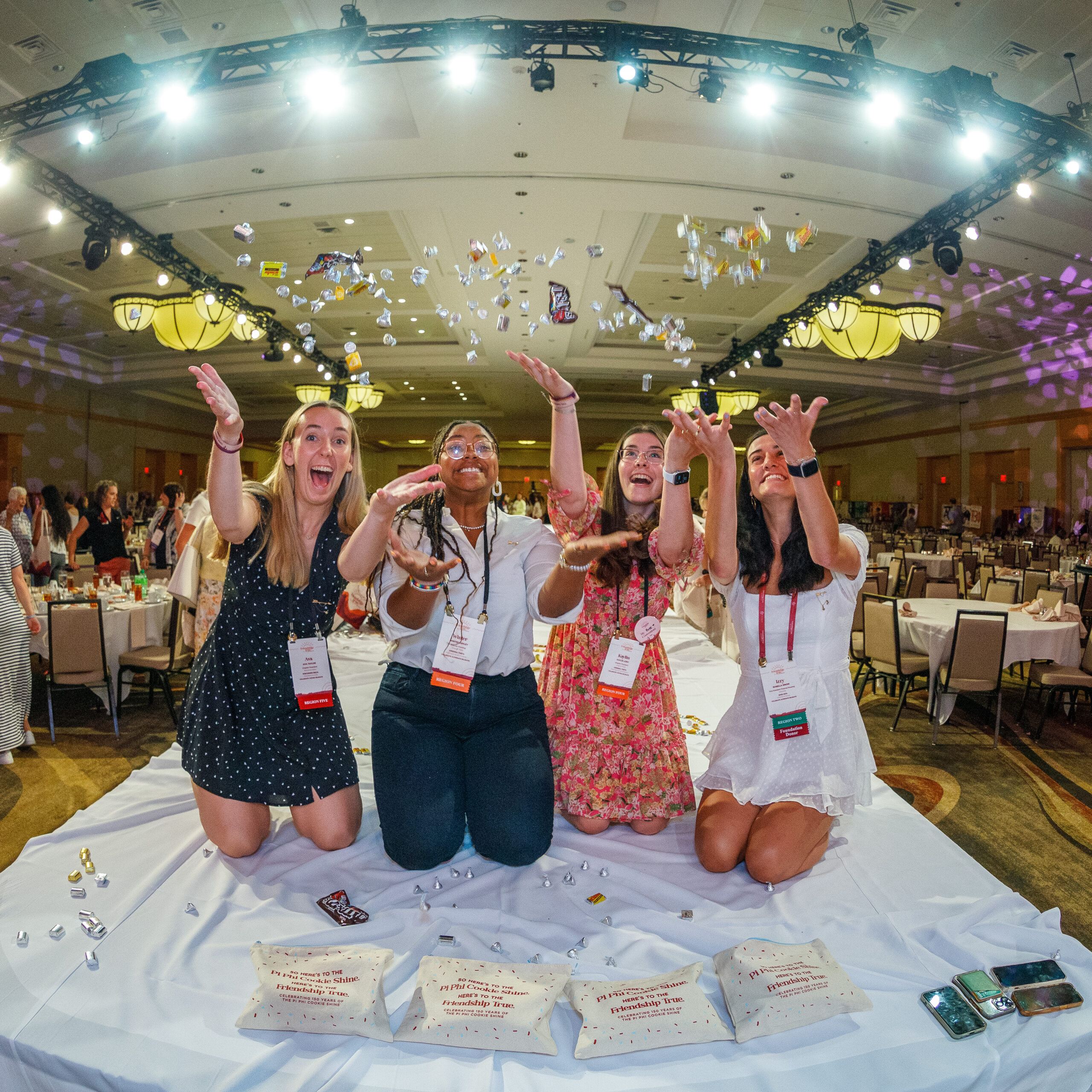 Four members kneeling on a table with a white tablecloth throwing candy in the air indoors at a conference with tables and chairs in the background