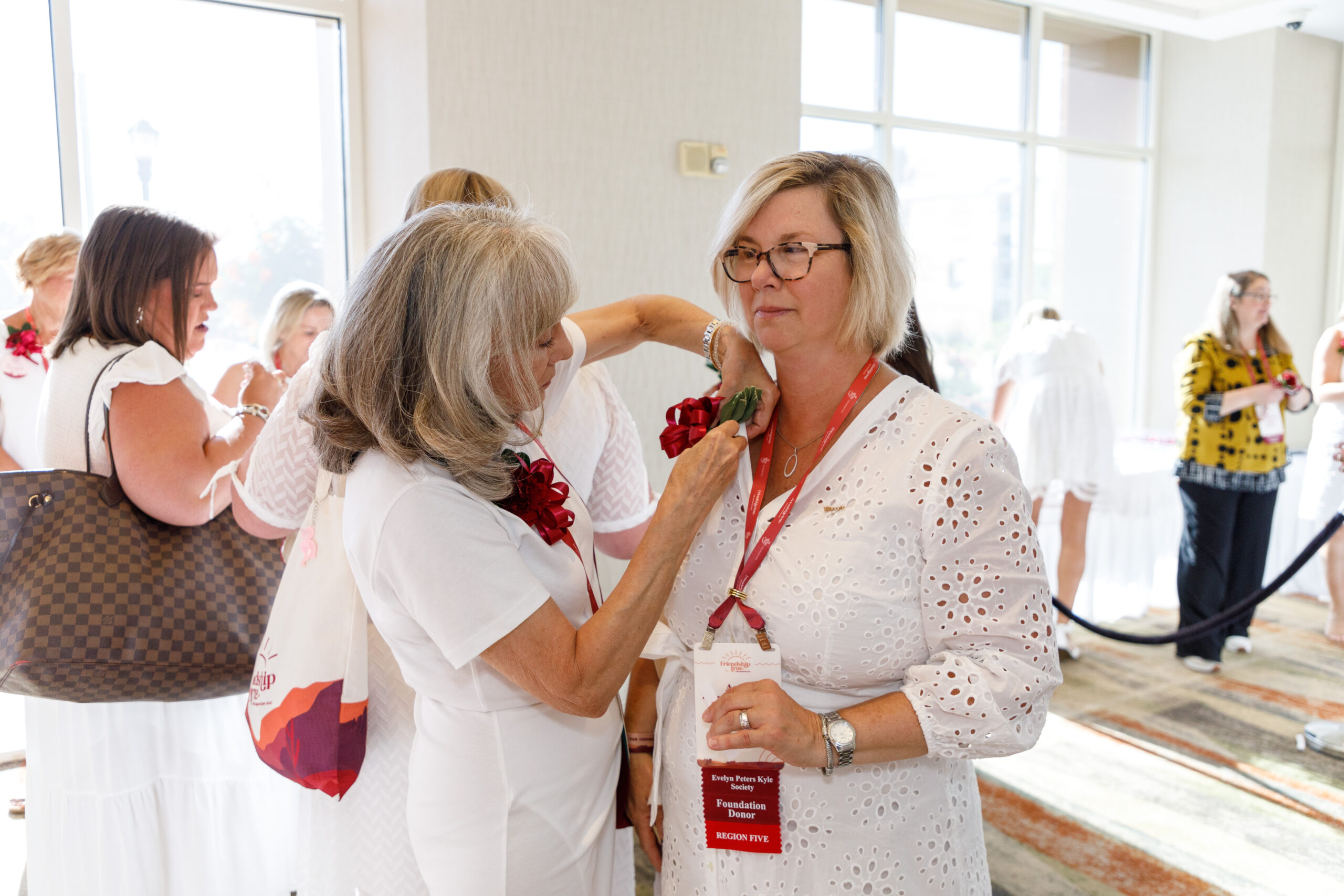 An alumnae member wearing a white dress pinning an alumnae member wearing a white dress with a red lanyard indoors with other people in the background