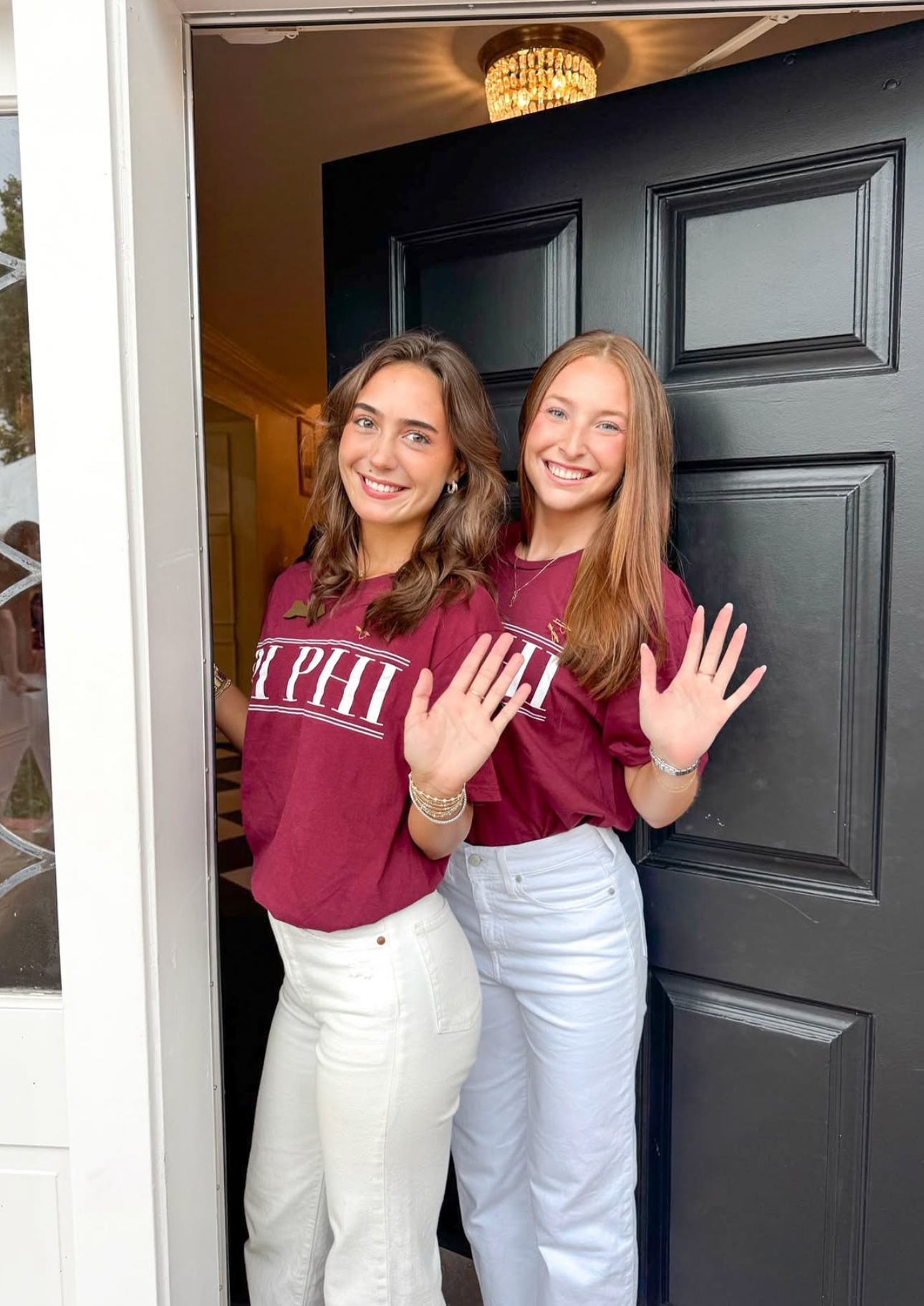 Two members wearing red Pi Beta Phi Greek letter shirts and white pants standing in front of a black door and white pillar