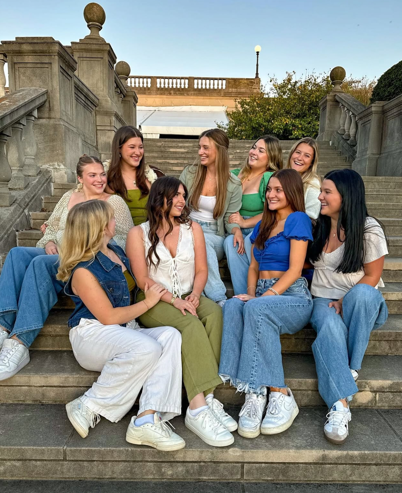 Nine member wearing jeans sitting outdoors on stone steps laughing and smiling at each other with evening light and a blue sky