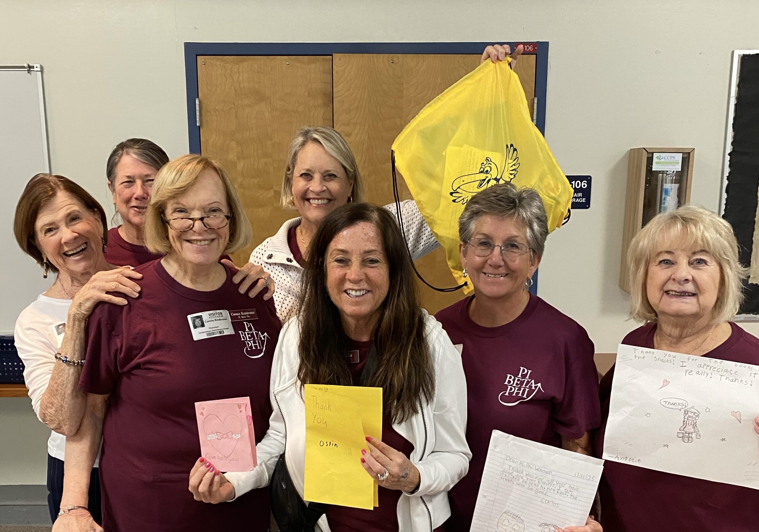 Seven alumnae members wearing wine colored Pi Beta Phi t-shirts standing in a school holding up yellow drawstring backpacks and white signs