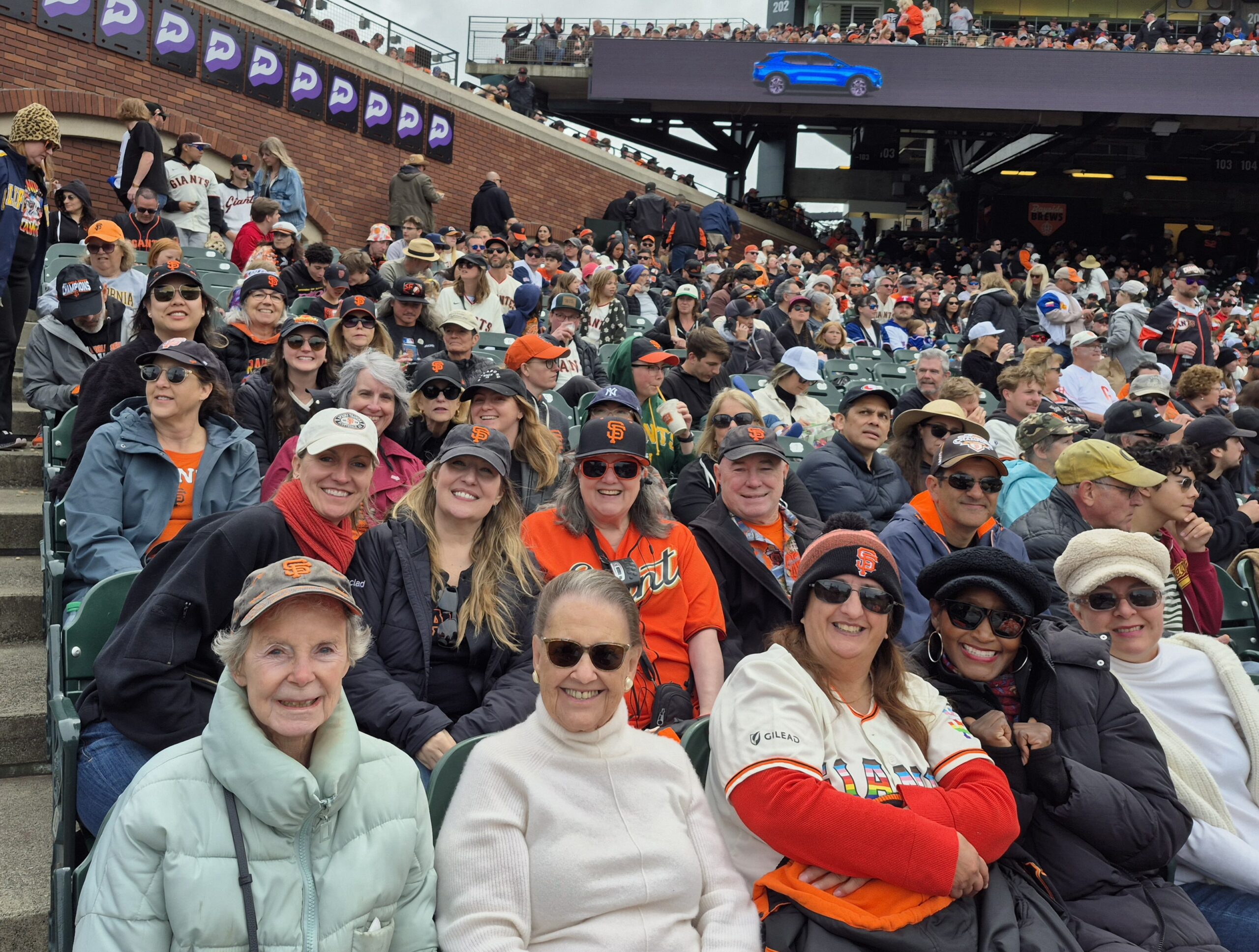 Several alumnae members wearing hats and sunglasses sitting in the stands of a baseball stadium with fans behind them