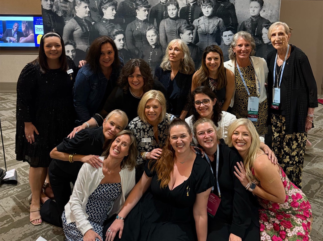 Large group of women happily posing in front of a large banner with a historic photo featured.