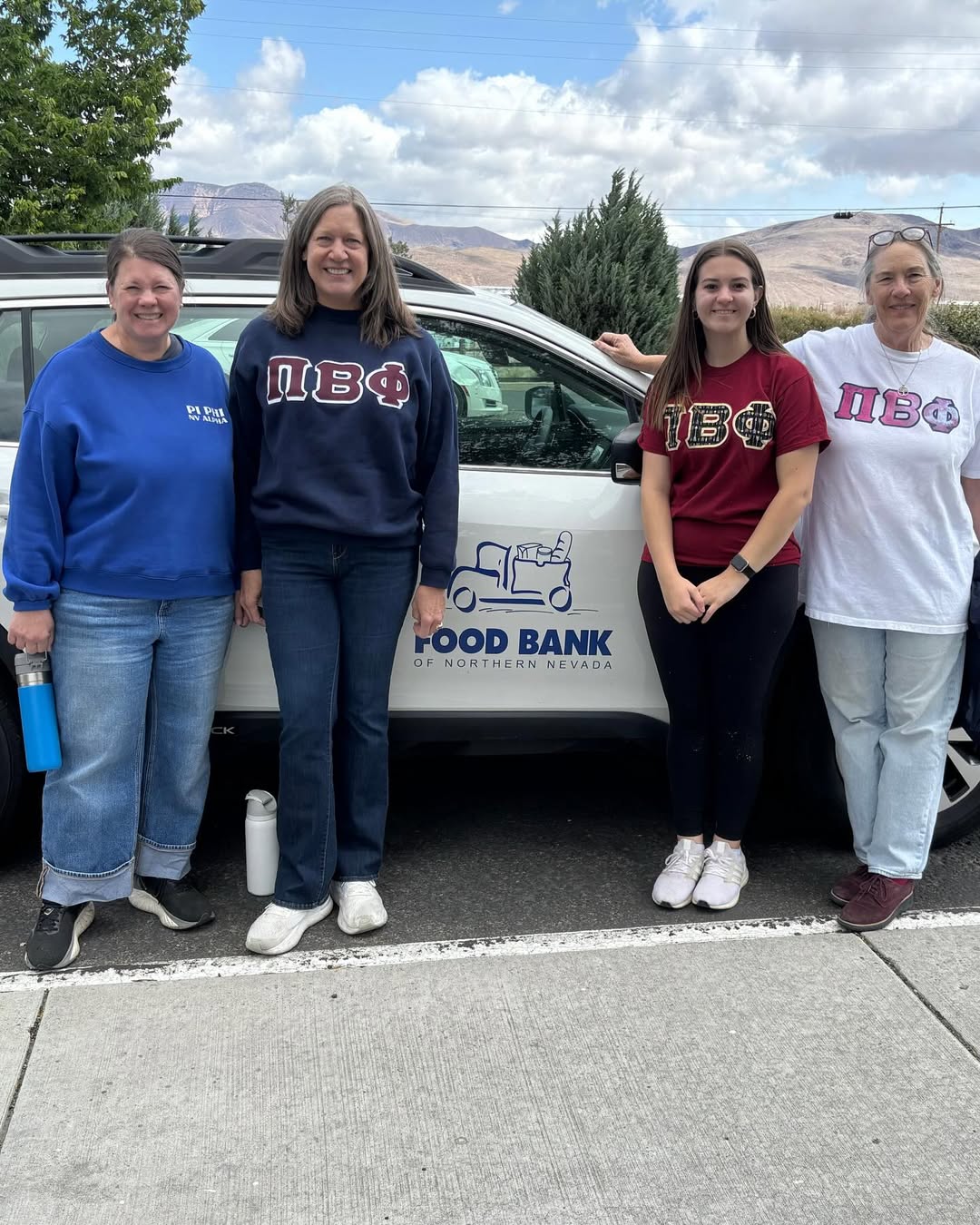 Four members wearing sweatshirts with Pi Beta Phi Greek letters standing outdoors in front of a white car with a food bank logo on the door