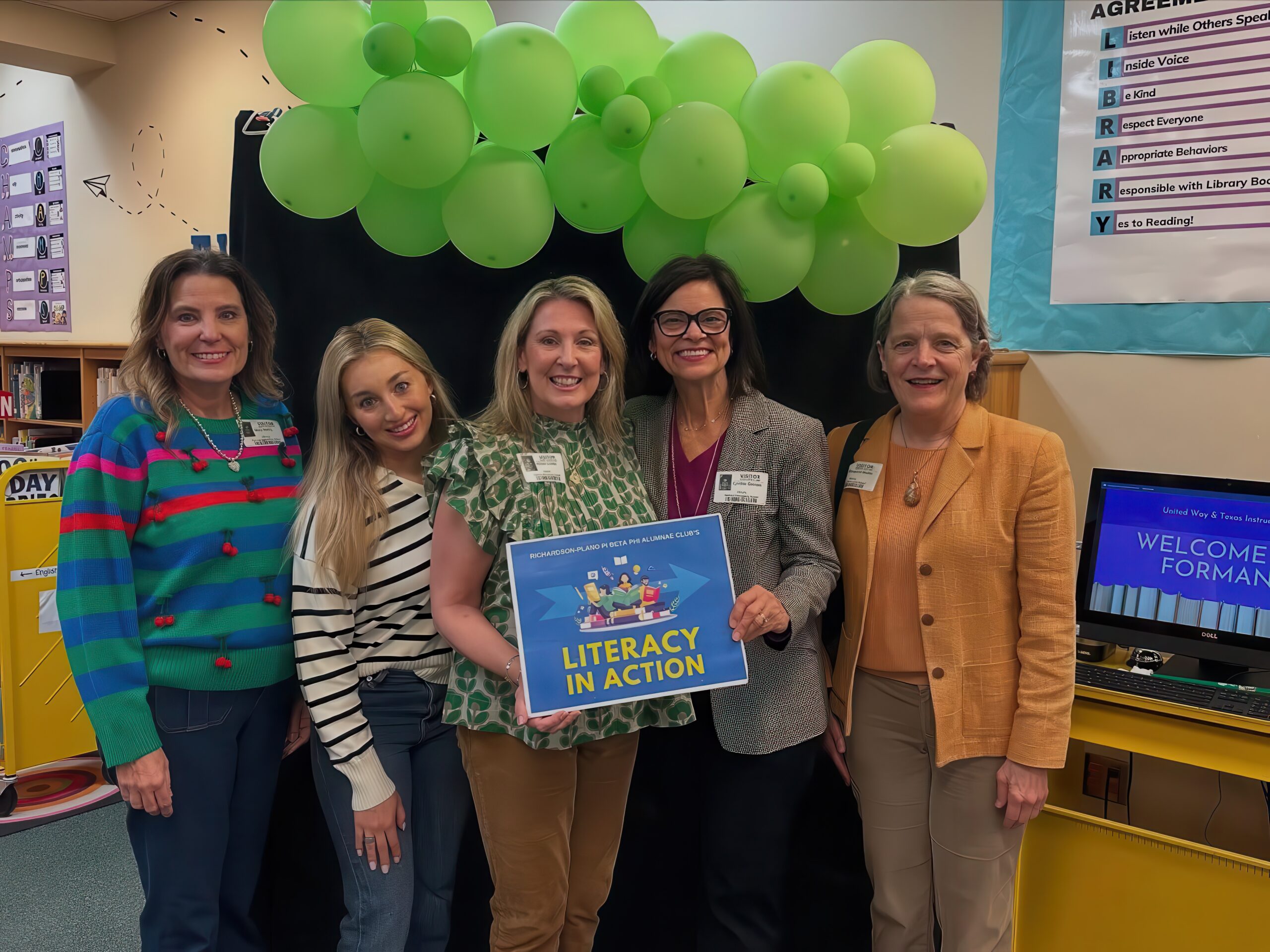 Five alumnae members standing in a classroom holding a blue sign that reads 