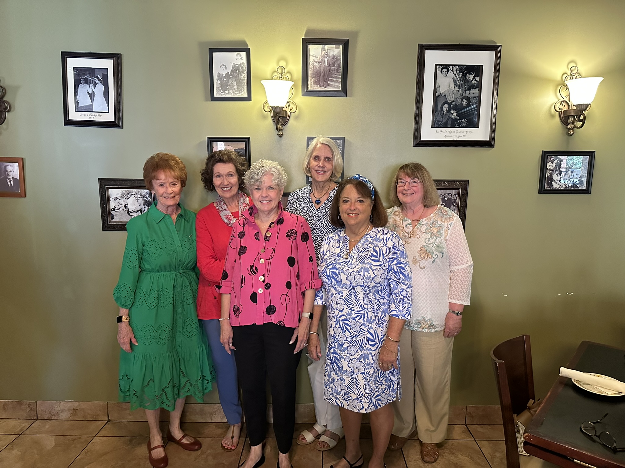 Six alumnae members wearing bright colored dresses standing in front of a beige wall with picture frames on it