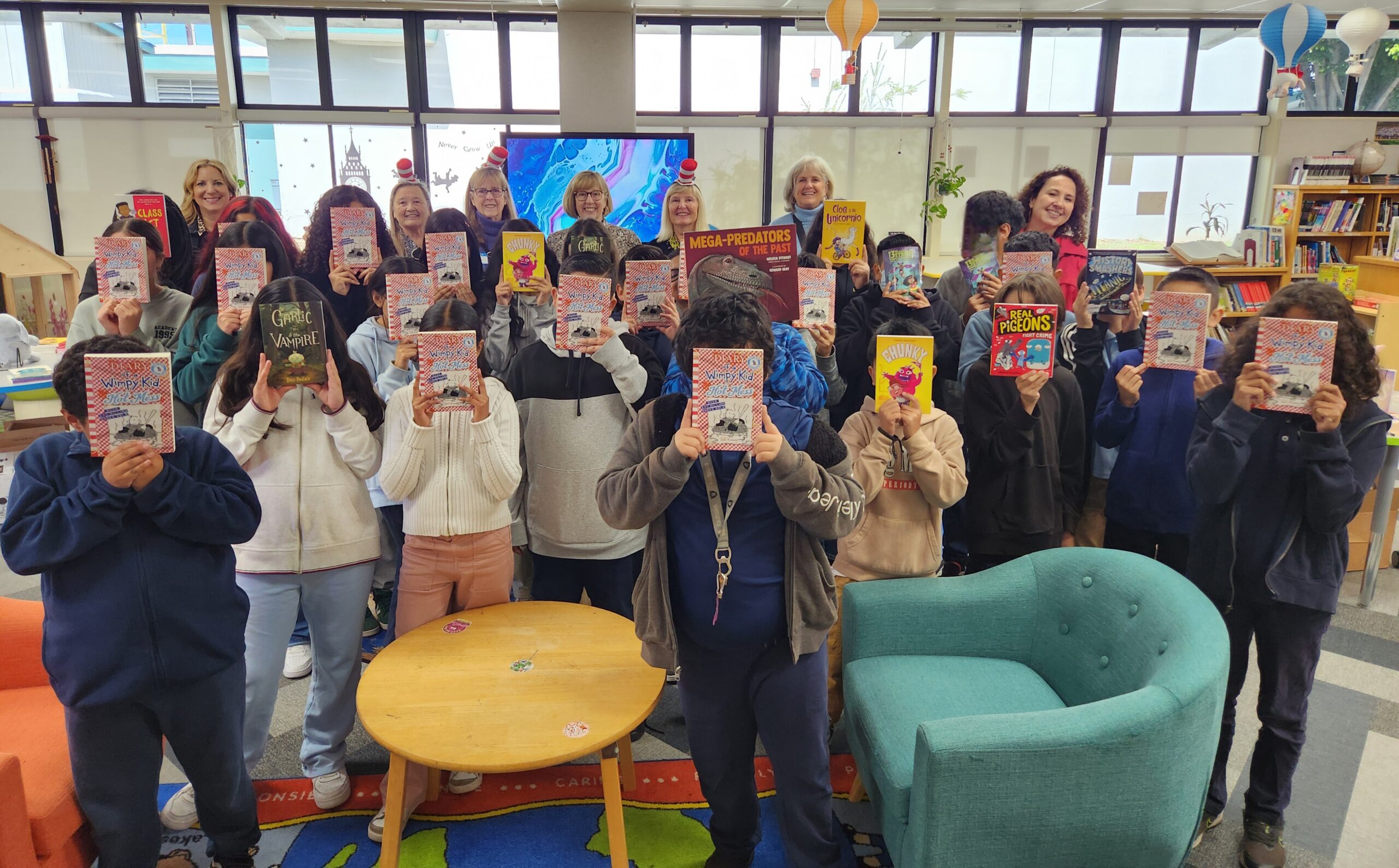 Alumnae members standing with children holding books in front of their faces in a classroom