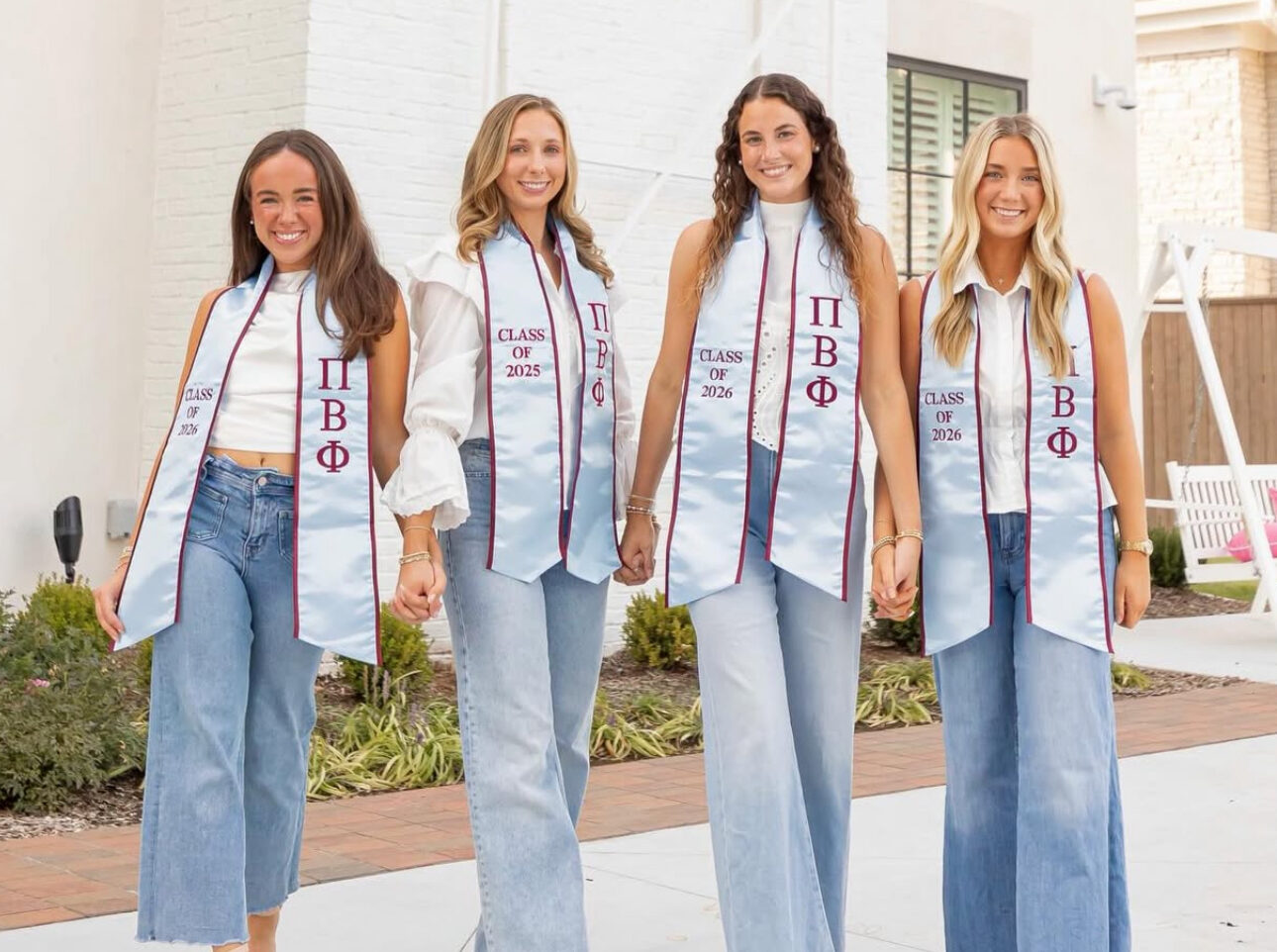 Four members wearing white shirt with jeans and Pi Beta Phi graduation stoles holding hands and standing outdoors in front of a large house