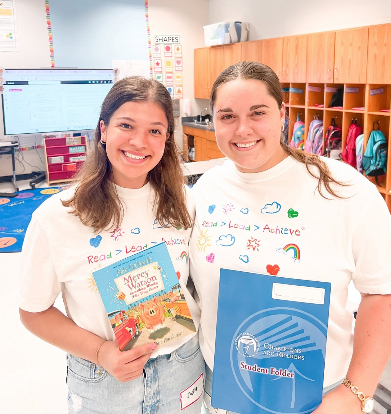 Two members wearing white t-shirts standing in a classroom in front of cubbies holding colorful books