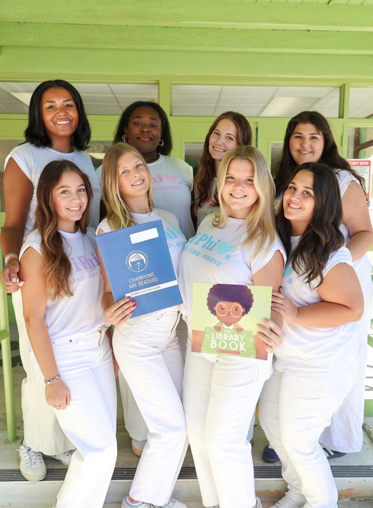 Eight members wearing white t-shirts and white pants standing in a classroom holding colorful books