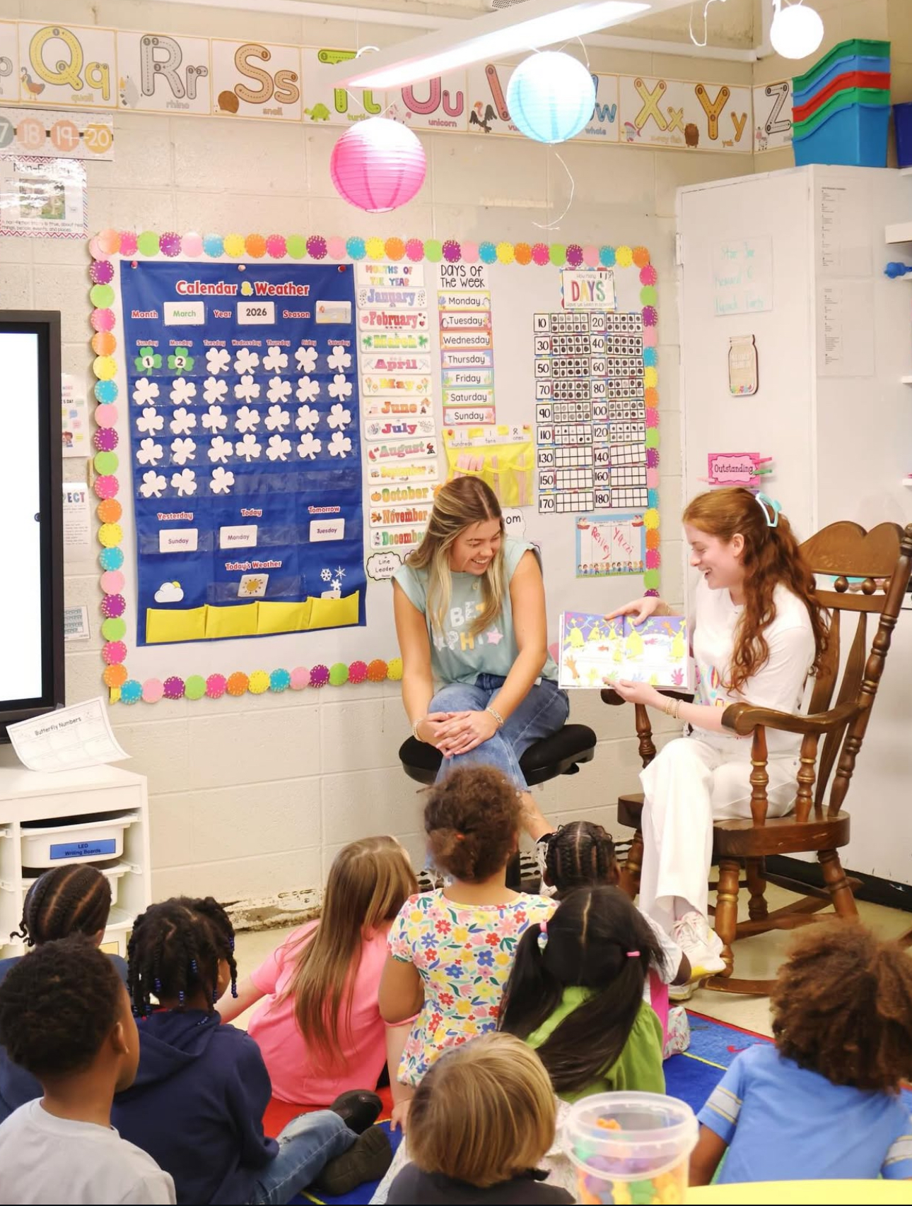 Two individuals sitting indoors reading a book to a classroom