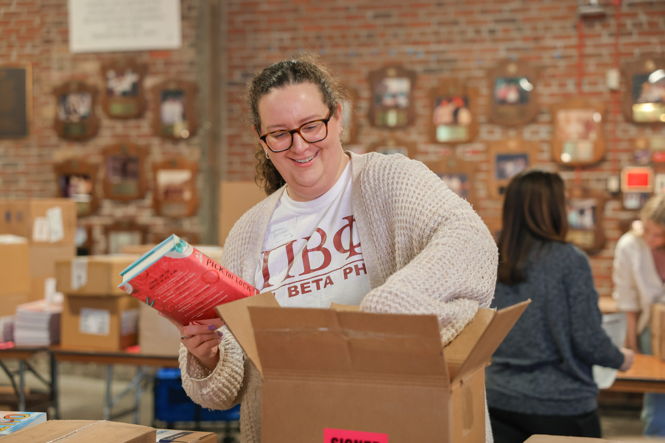 One member wearing a white t-shirt with Pi Beta Phi Greek letters and a beige cardigan putting red books into a box in front of a brick wall with picture frames