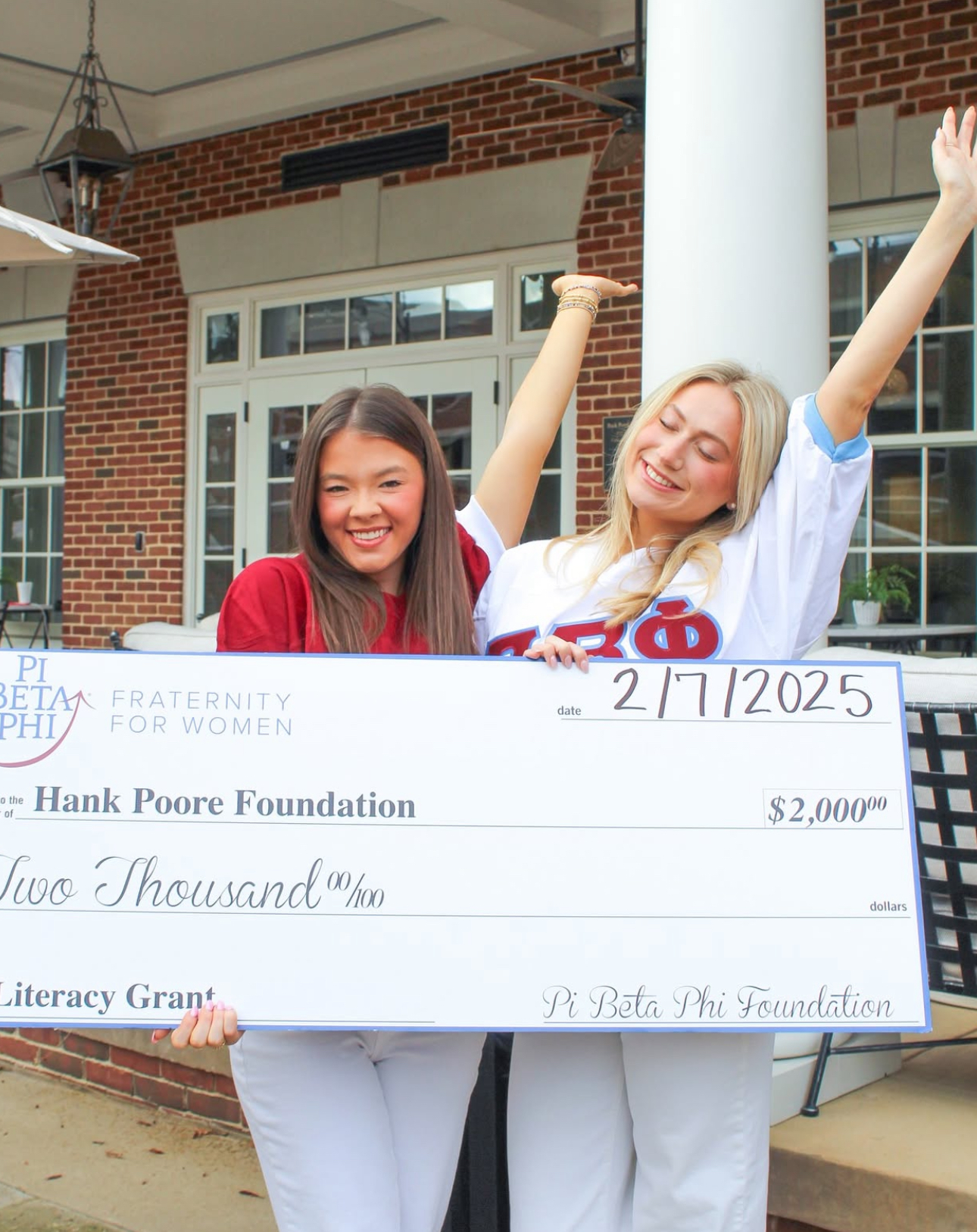 Two members standing outside with their arms raised holding a large check in front of a brick chapter facility with a white pillar