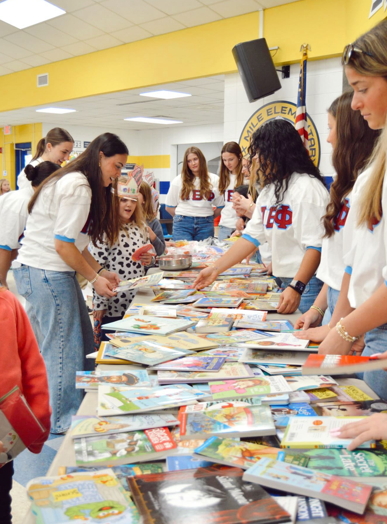 A group of members wearing white t-shirt with Pi Beta Phi Greek letters sorting colorful books at a narrow table inside a school