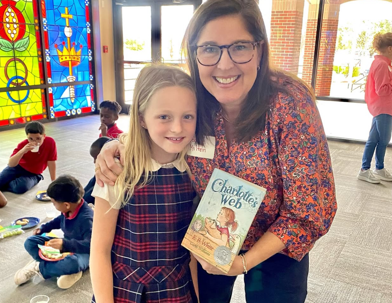One member wearing a red blouse posing with a young student in a plaid dress hold the book "Charlotte's Web" in a classroom