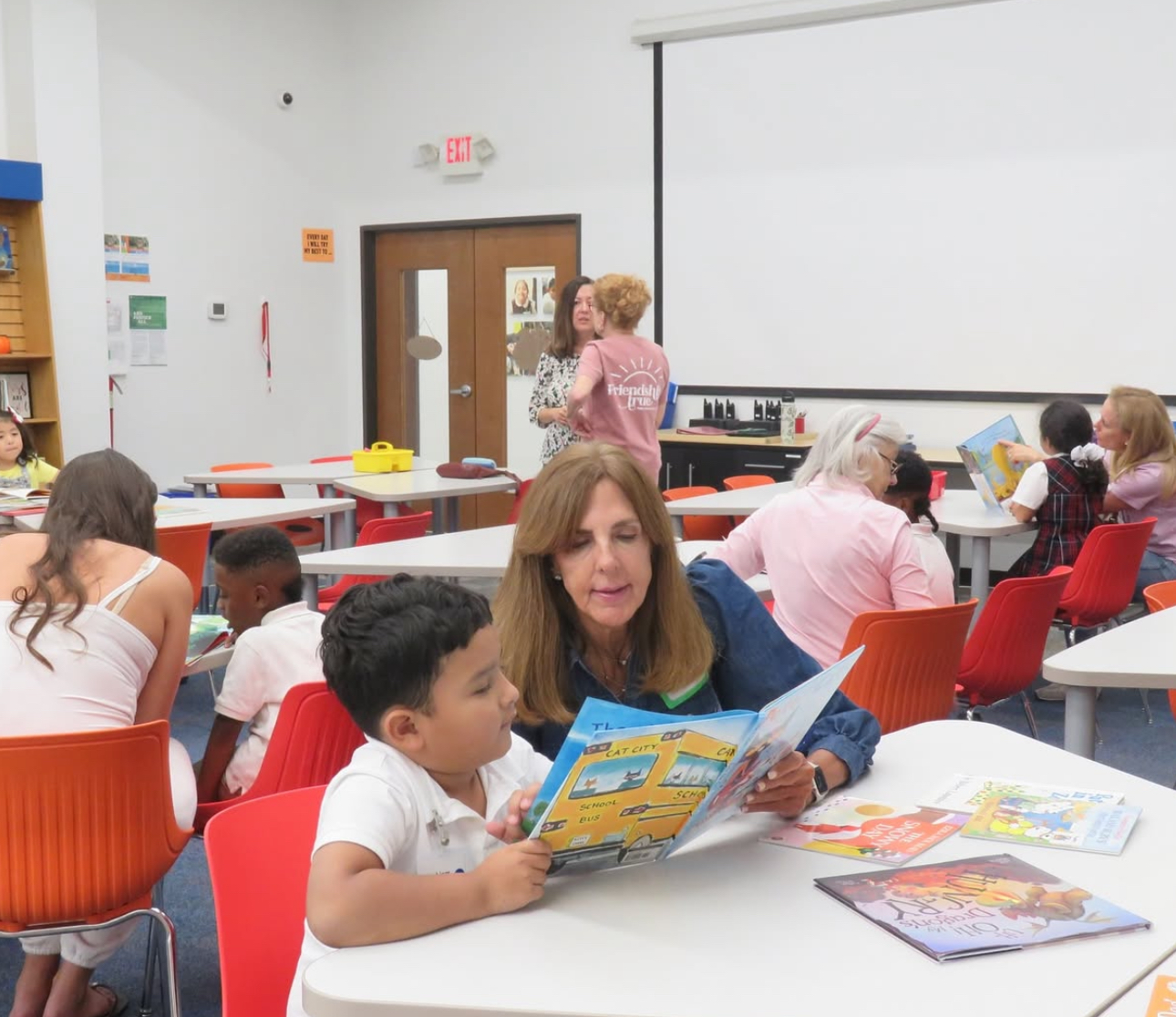 One member reading a blue book to a child sitting down at a table in a classroom with other students in the background