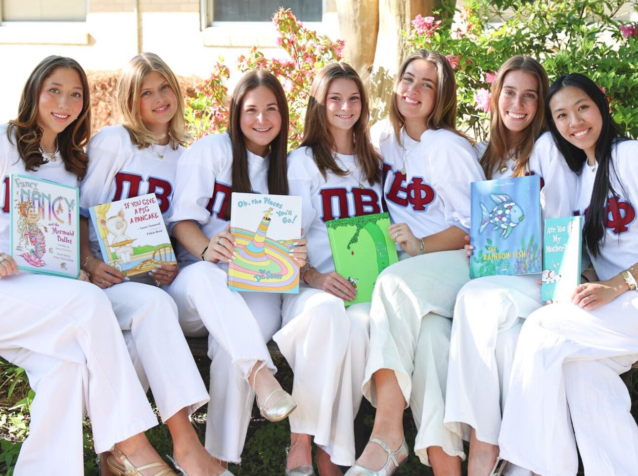 Seven members wearing white t-shirts with Pi Beta Phi Green letters and white pants sitting outdoors holding four Dr. Suess books