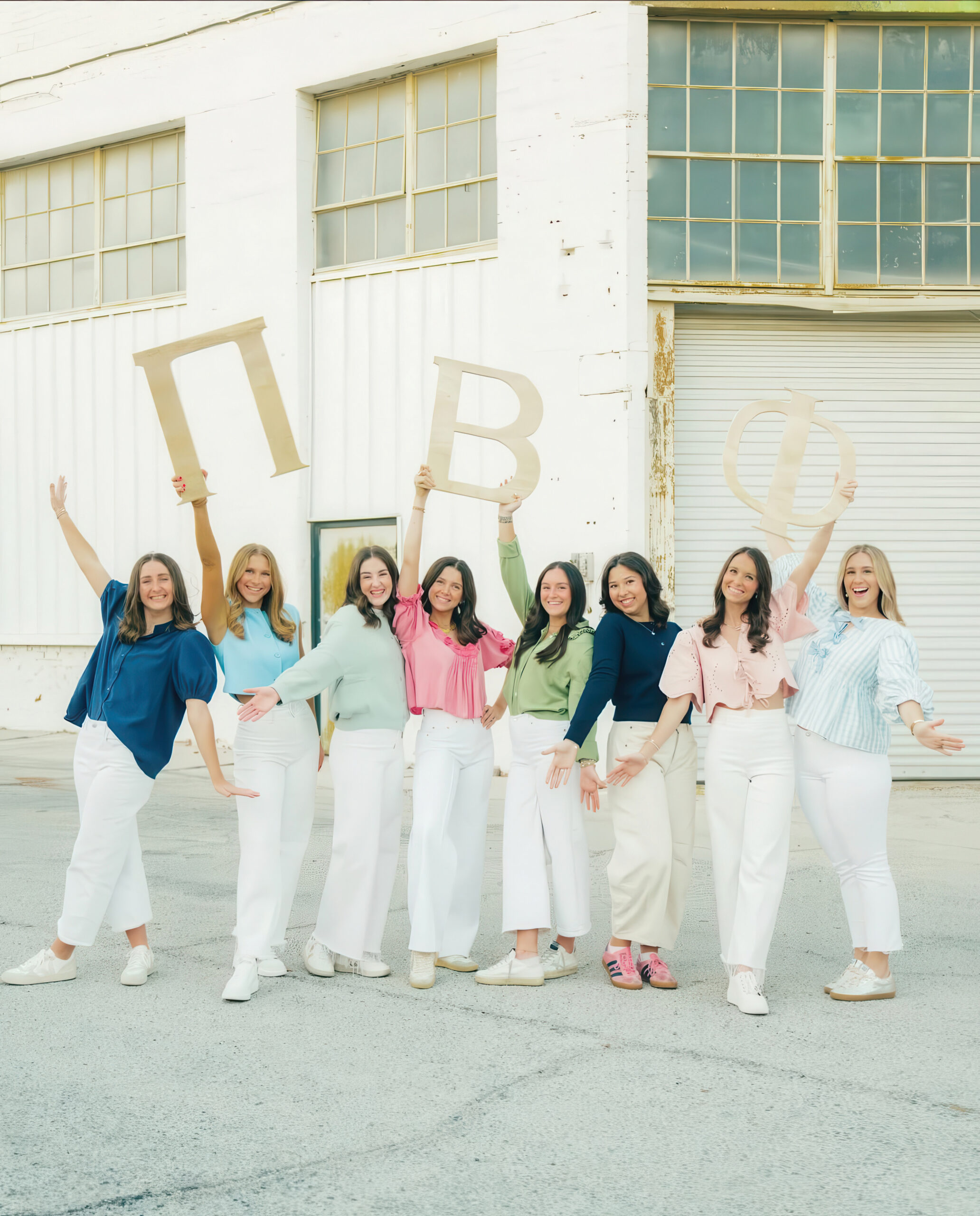 Eight members wearing colorful shirts and white pants standing outdoors in front of a while building holding up large Pi Beta Phi Greek letters
