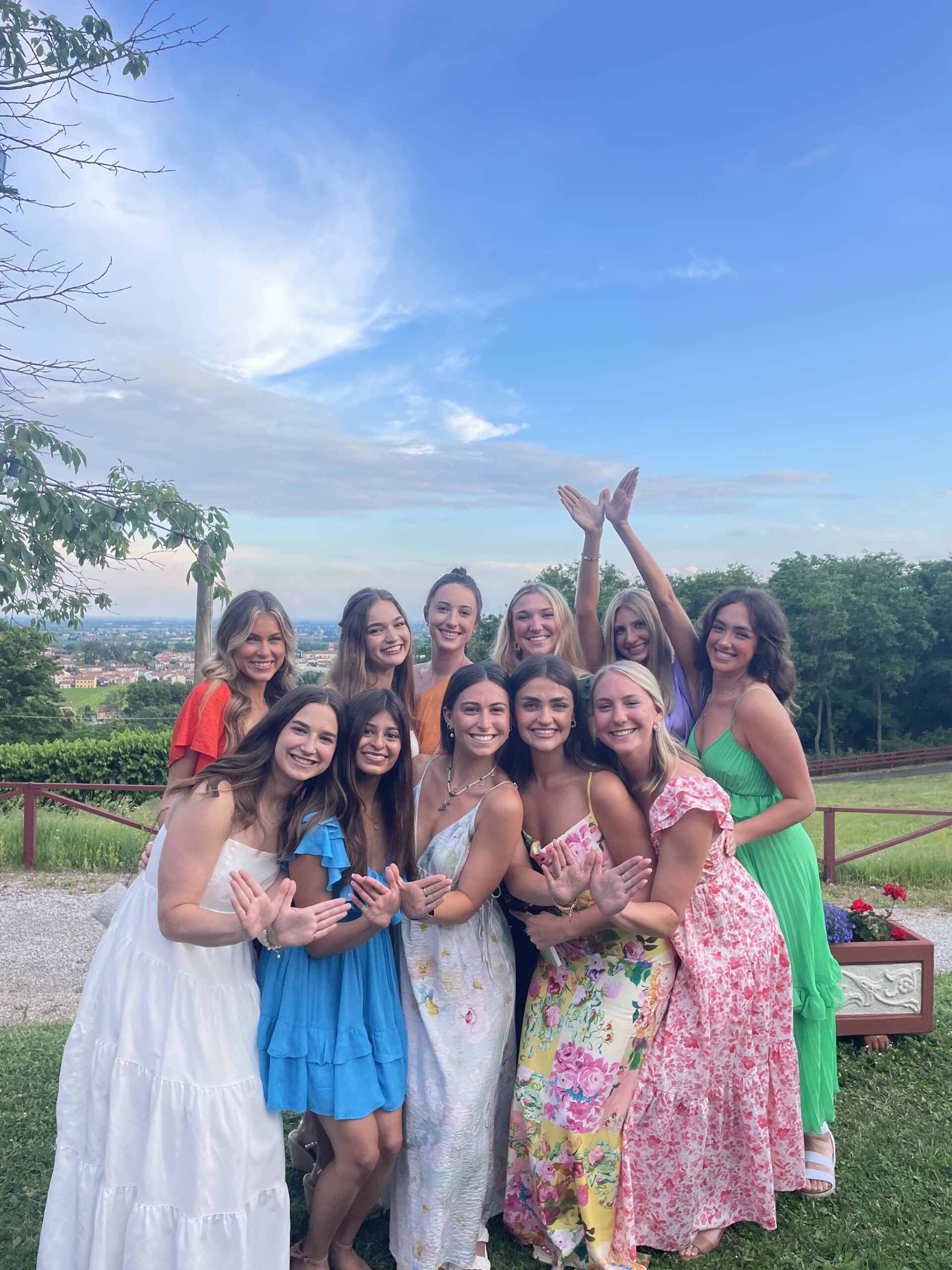 Several members wearing colorful dresses standing outside in front of green trees with one member holding up angel wings in the background