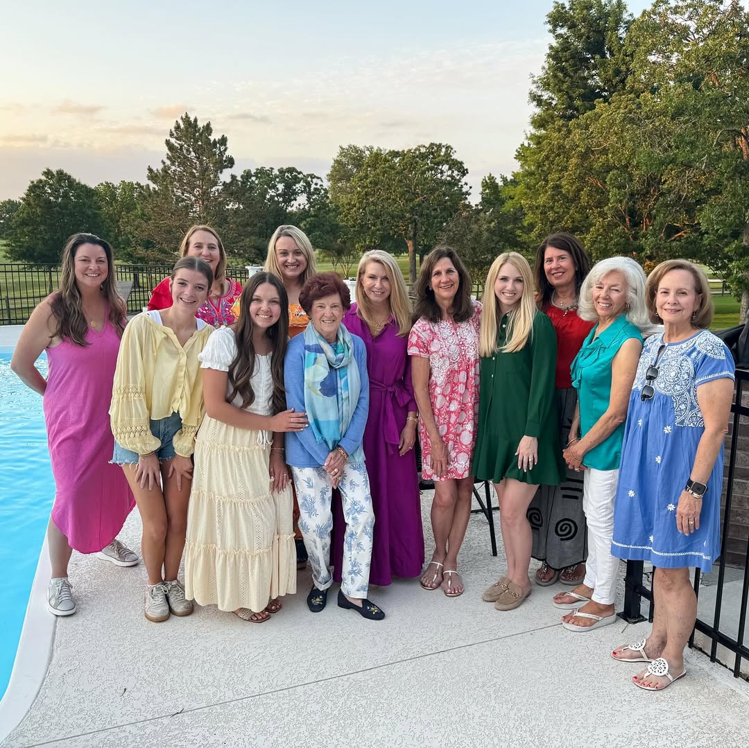 Several alumnae members wearing colorful dresses standing outside next to a pool with a sunset and green trees in the background