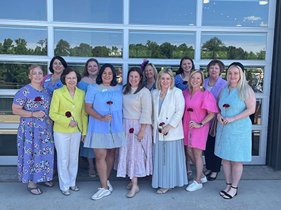 Several alumnae members wearing colorful dresses and holding red carnations standing outdoors in front of a large window