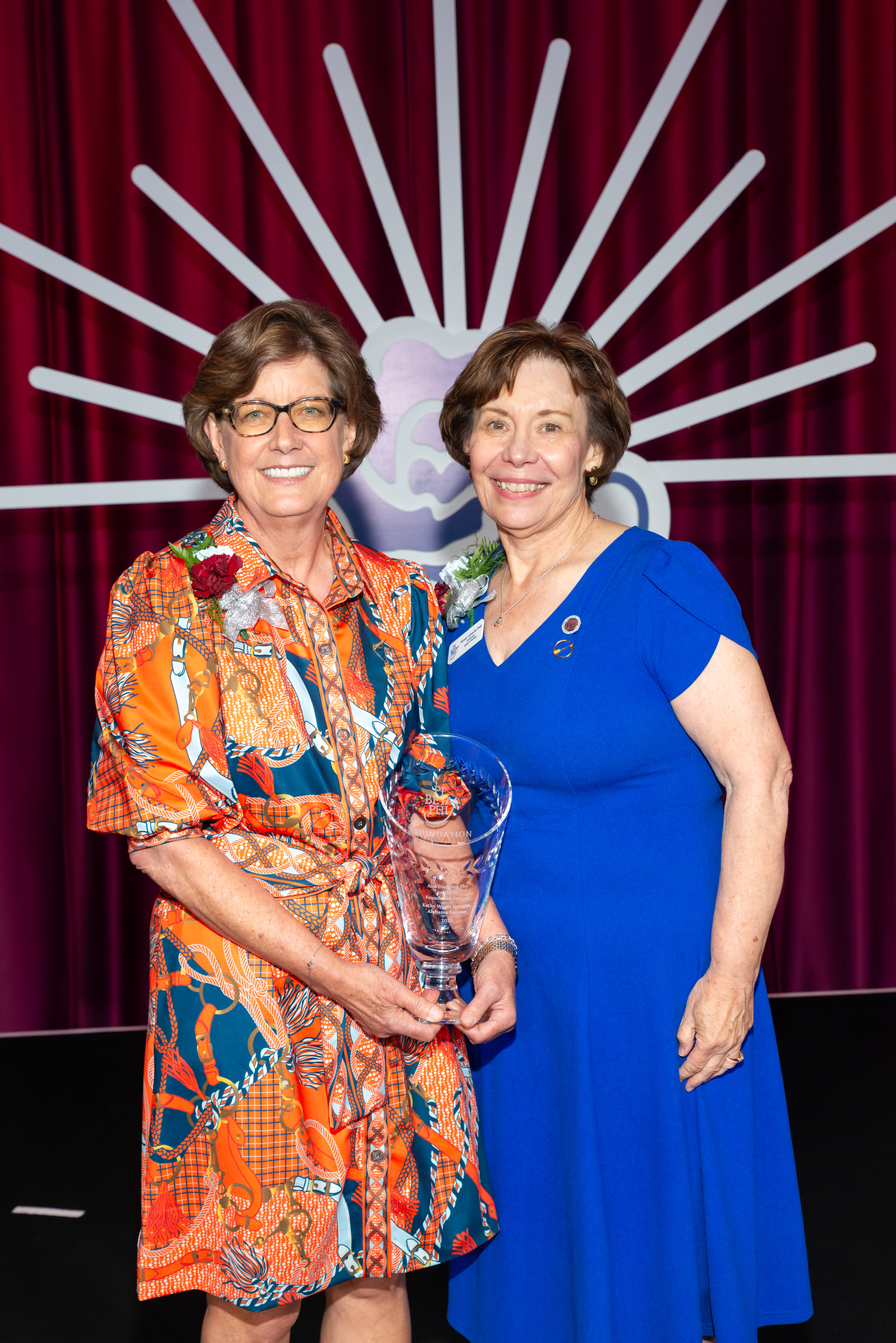 Two alumnae members wearing colorful dresses standing on stage with an award in front of a sunburst background