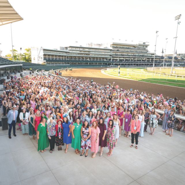 Hundreds of Pi Beta Phi alumnae posing for a large group photo on the terrace at Churchill Downs racetrack, wearing colorful dresses and decorative hats