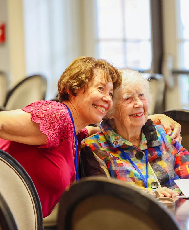Two Pi Beta Phi alumnae smiling and hugging at an indoor reunion banquet, wearing a pink lace top and colorful patterned jacket with convention lanyards