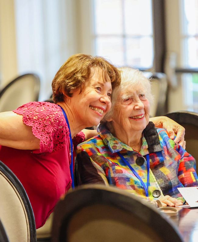 Two Pi Beta Phi alumnae smiling and hugging at an indoor reunion event, wearing conference lanyards and seated at a banquet table
