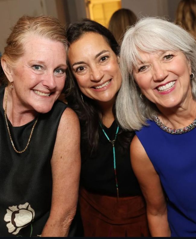 Three Pi Beta Phi alumnae smiling together at an indoor evening event, wearing black, brown, and royal blue outfits with statement necklaces