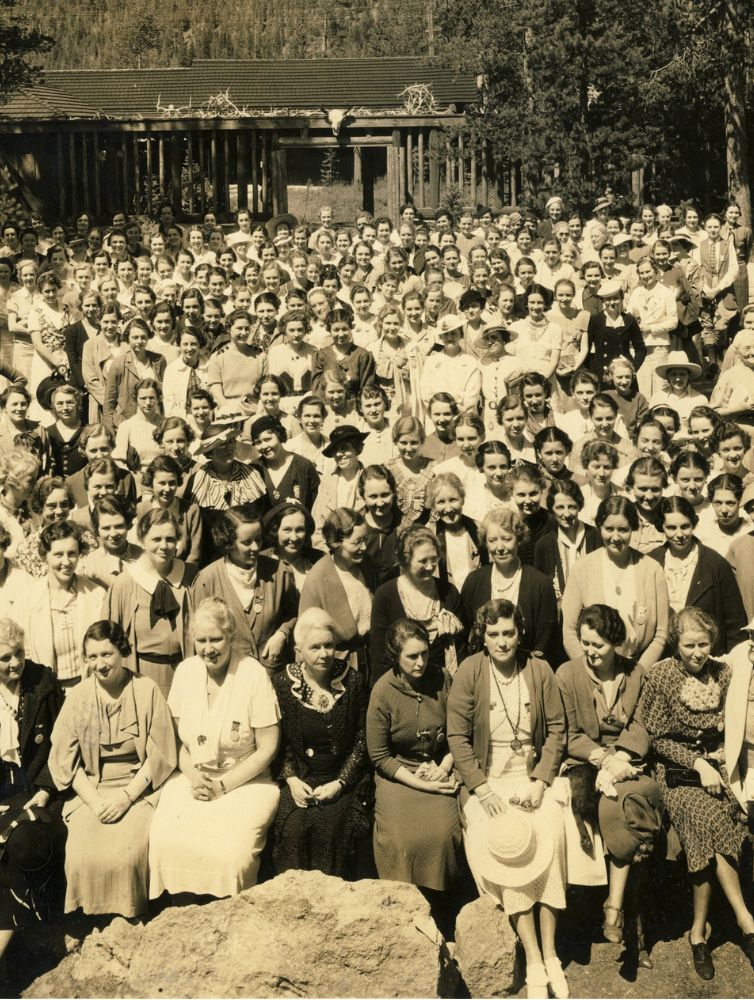 Sepia-toned historical photograph of a large group of Pi Beta Phi members gathered outdoors in front of a rustic lodge surrounded by pine trees