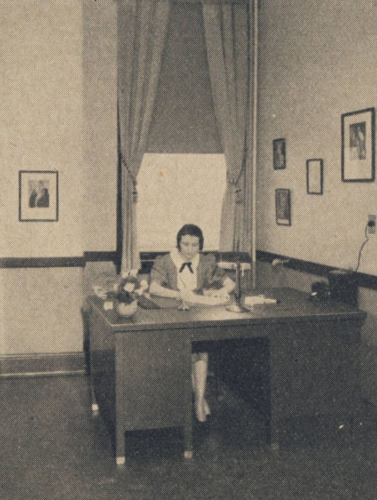 Black and white historical photograph of a Pi Beta Phi staff member working at a desk in a formal office with framed portraits on the walls