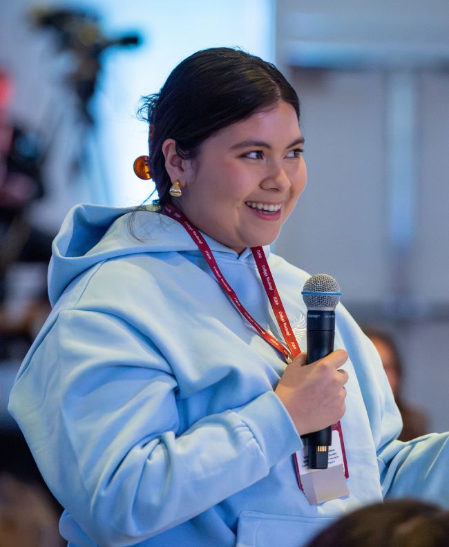 Pi Beta Phi member smiling and speaking into a microphone at an indoor conference, wearing a light blue hoodie and event lanyard