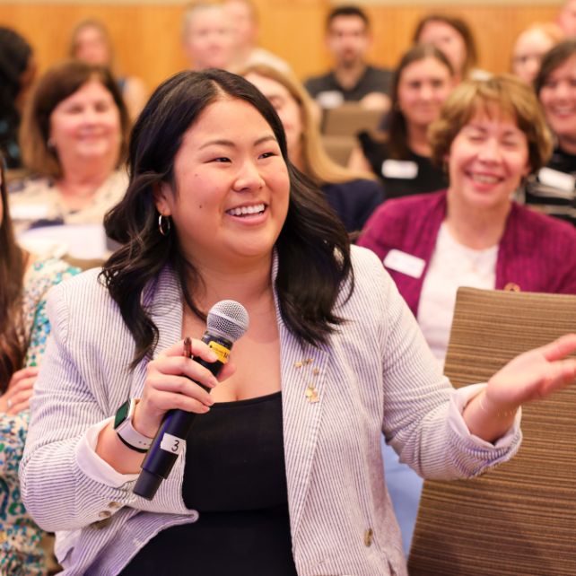 Pi Beta Phi member smiling and speaking into a microphone during an indoor officers workshop session, with a room full of engaged attendees laughing and listening behind her