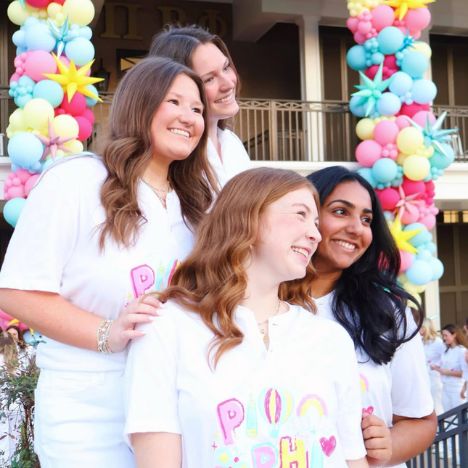 Four Pi Beta Phi members smiling together outdoors in front of a colorful balloon arch, wearing white Pi Beta Phi t-shirts at a bid day celebration