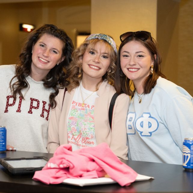 Pi Beta Phi members smiling together outdoors at a College Weekend event, wearing casual summer outfits with name badge lanyards
