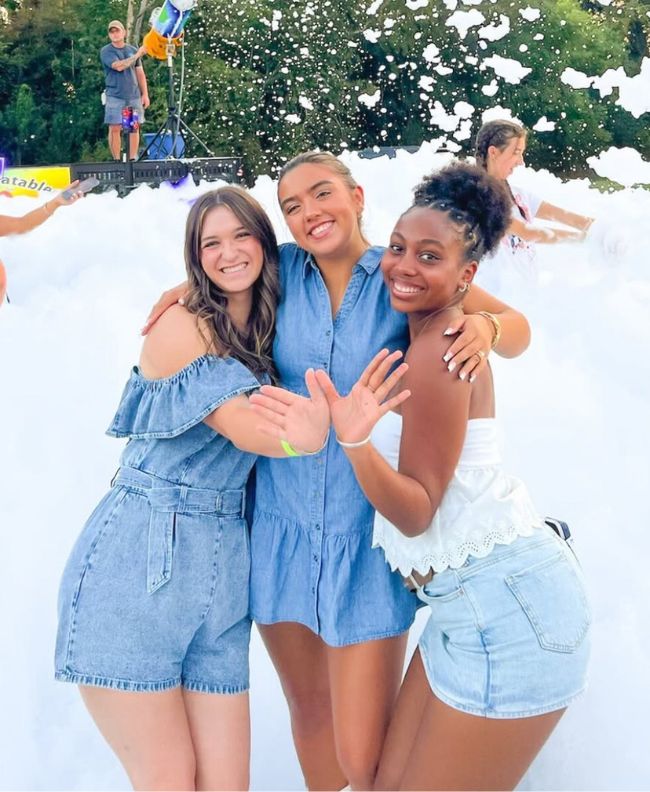 Three Pi Beta Phi members smiling and posing together at an outdoor foam party, wearing denim outfits surrounded by foam with trees in the background