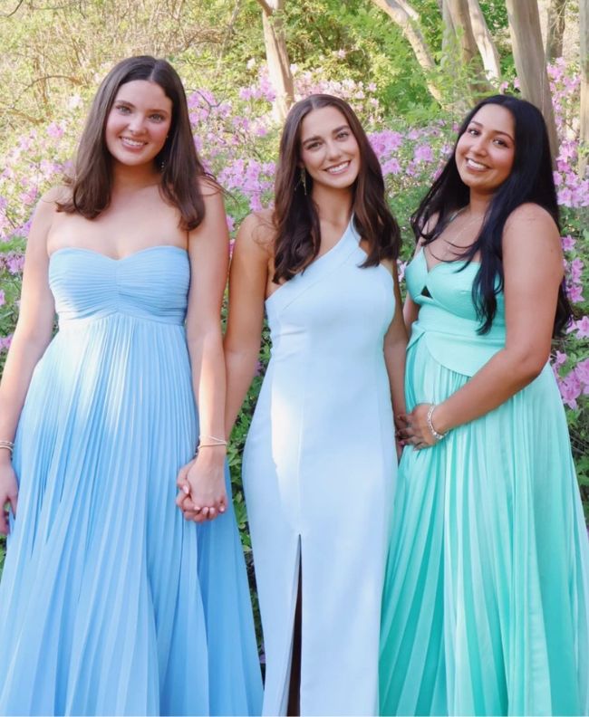 Three Pi Beta Phi members smiling together in a spring garden with blooming pink azaleas, wearing blue, white, and teal formal gowns