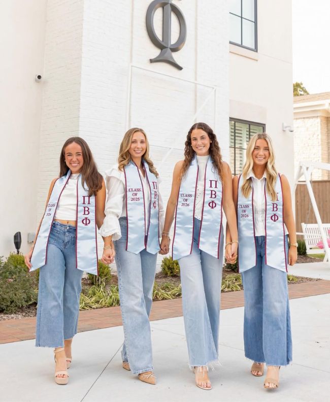 Four Pi Beta Phi members smiling and walking together in front of the chapter house wearing Pi Beta Phi graduation stoles for the classes of 2025 and 2026 with jeans and heels