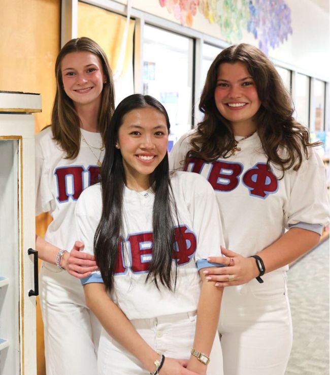Three Pi Beta Phi members smiling together indoors wearing white Greek letter jerseys
