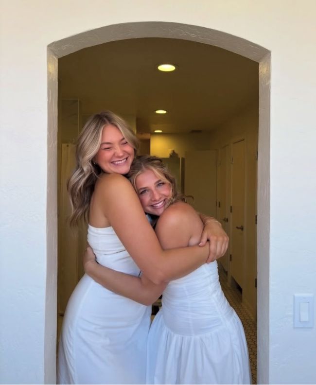 Two Pi Beta Phi members laughing and hugging tightly in an arched doorway, both wearing white dresses