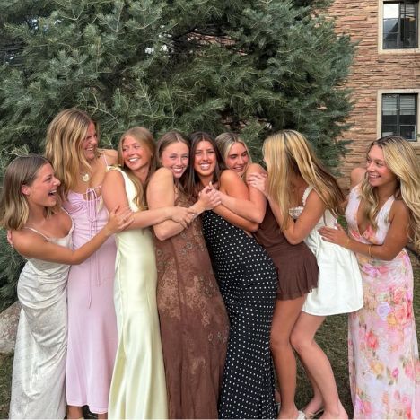Eight Pi Beta Phi members laughing and hugging together outdoors in front of a pine tree, wearing colorful summer dresses at a formal event