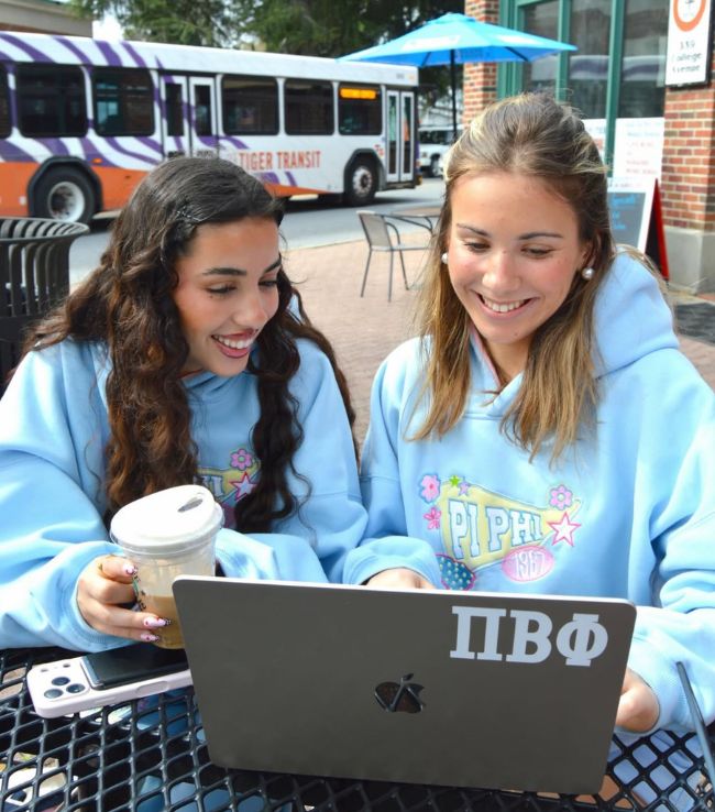 Two Pi Beta Phi members in light blue hoodies smiling at a laptop with a Pi Beta Phi sticker at an outdoor campus cafe