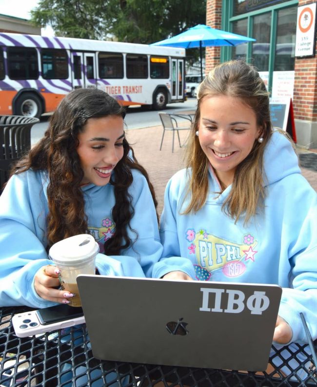 Two Pi Beta Phi members smiling at a laptop with a Pi Beta Phi sticker at an outdoor campus cafe table, wearing light blue Pi Phi hoodies, one holding a coffee cup