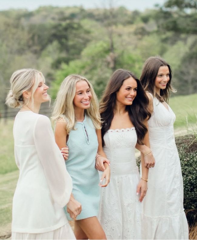 Four Pi Beta Phi members laughing together outdoors in a green field, wearing white, blue, and cream dresses with pine trees in the background