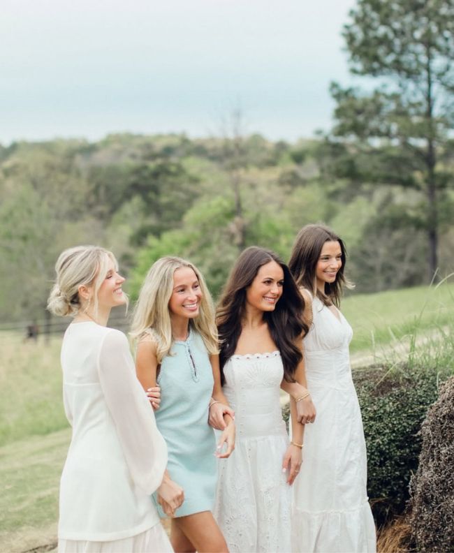 Four Pi Beta Phi members laughing together outdoors in a green rural landscape with pine trees, wearing white, light blue, and cream dresses