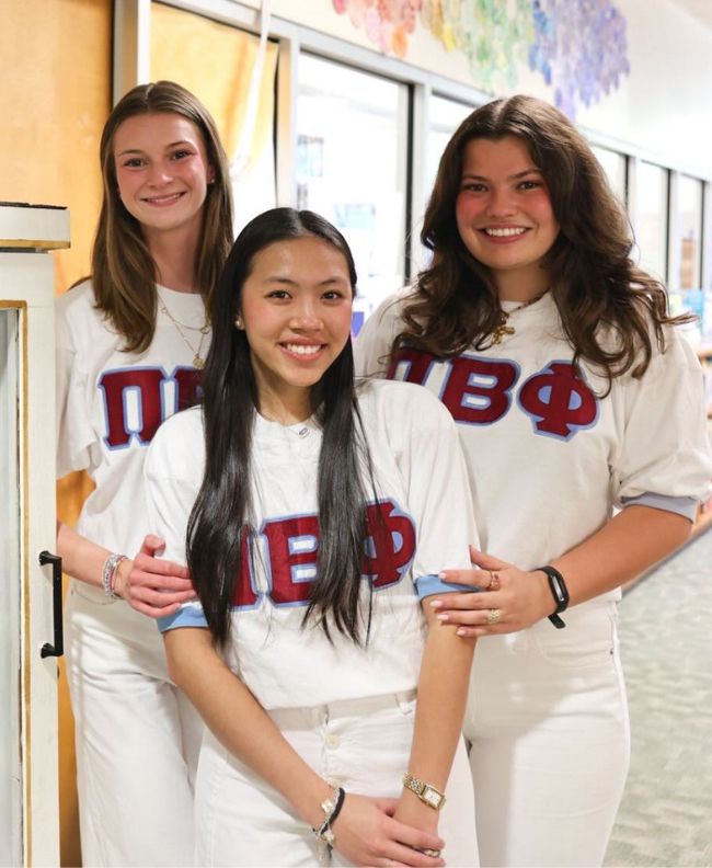 Three Pi Beta Phi members smiling next to a little free library filled with children's books, wearing white Pi Beta Phi Greek letter jerseys inside a school hallway