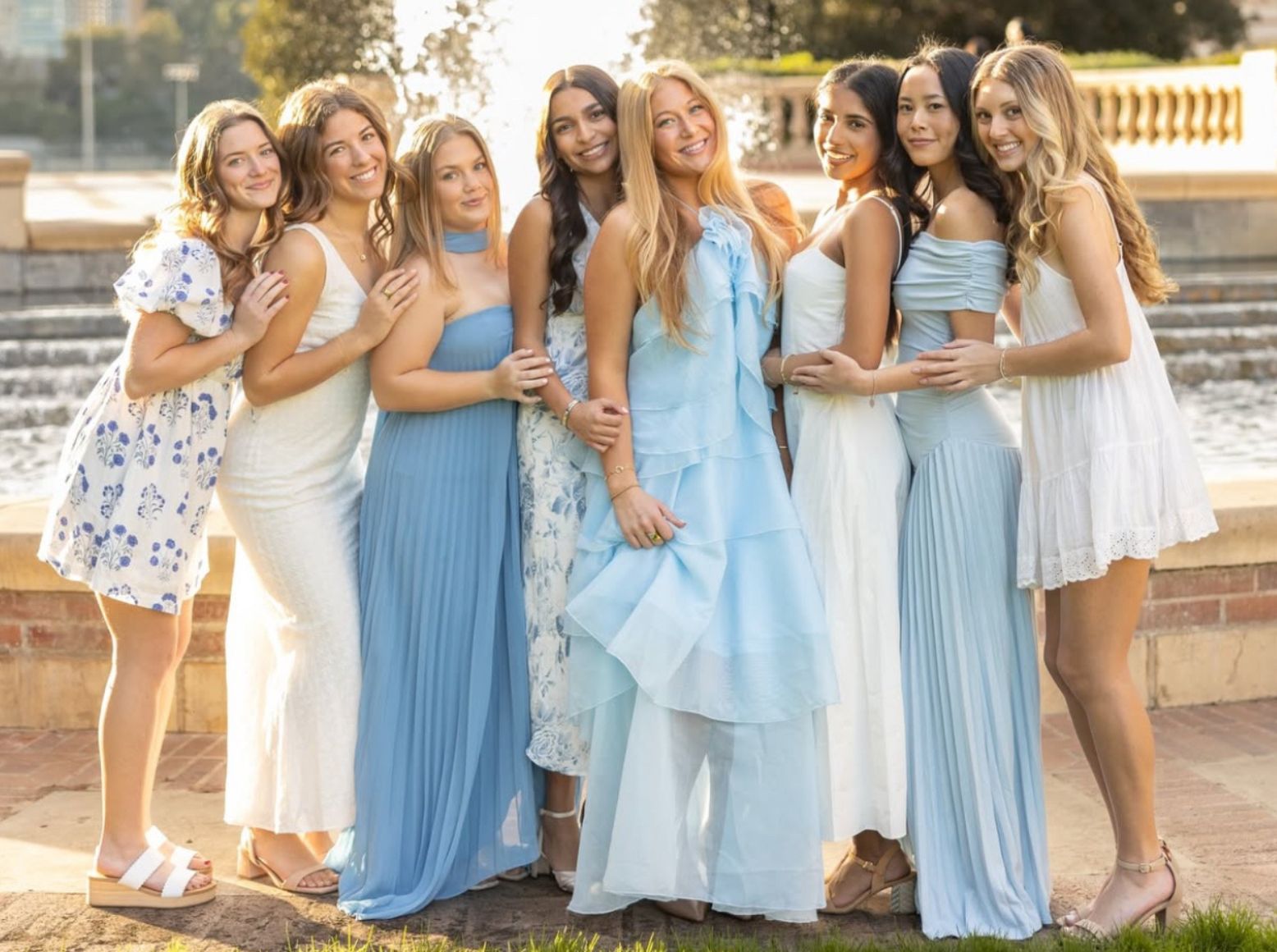 Eight Pi Beta Phi members smiling together in front of an outdoor fountain at golden hour, wearing coordinating blue and white dresses
